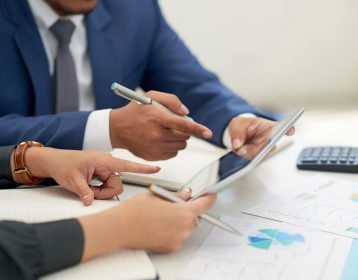 unrecognizable-business-people-sitting-at-meeting-with-charts-looking-and-pointing-at-tablet