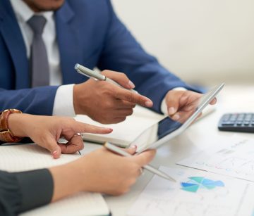 unrecognizable-business-people-sitting-at-meeting-with-charts-looking-and-pointing-at-tablet